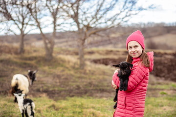 Cute girl in red jacket and beanie holding black baby goat on farm. Rural lifestyle, childhood, animal care countryside setting
