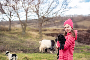 Cute girl in red jacket and beanie holding black baby goat on farm. Rural lifestyle, childhood, animal care countryside setting