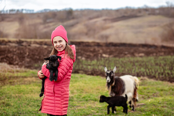 Cute girl in red jacket and beanie holding black baby goat on farm. Rural lifestyle, childhood, animal care countryside setting