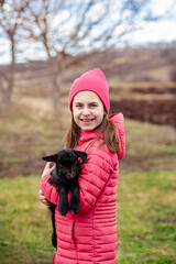 Cute girl in red jacket and beanie holding black baby goat on farm. Rural lifestyle, childhood, animal care countryside setting