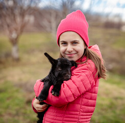 Cute girl in red jacket and beanie holding black baby goat on farm. Rural lifestyle, childhood, animal care countryside setting