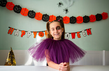 Girl Smiling in Purple Halloween Costume. Smiling girl in purple tulle costume with Halloween makeup, sitting in decorated room with pumpkins, garlands, spiders, and witch hat.