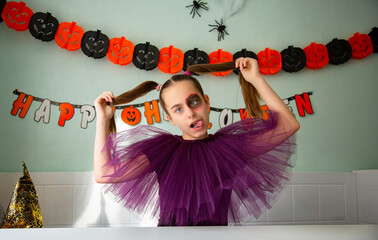 Girl Smiling in Purple Halloween Costume. Smiling girl in purple tulle costume with Halloween makeup, sitting in decorated room with pumpkins, garlands, spiders, and witch hat.