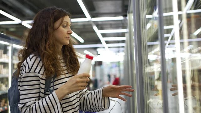 Young woman with curly hair shopping in a grocery store, opening a refrigerator door to choose a bottle of kefir or milk and carefully reading the label before making a healthy food choice