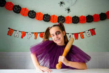 Smiling girl in Halloween makeup, wearing black costume and holding toy axe, sitting in festively decorated room pumpkins