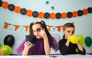 Two kids getting ready for Halloween. One girl applies makeup while boy inflates balloon.