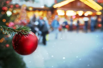 Rote Christbaumkugel, Weihnachtsschmuck, Tannenzweig auf Weihnachtsmarkt, Christkindlmarkt vor Verkaufsstand, Hütten, Schneefall, Schneeflocken, Lichtern, verschwommener Hintergrund im kalten Winter. © Barbara-Maria Damrau