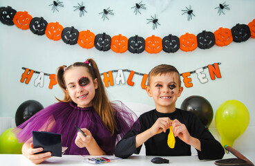 Two kids getting ready for Halloween. One girl applies makeup while boy inflates balloon.