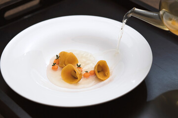 Gourmet orange tortellini with garnish being finished with a clear broth poured from a glass teapot onto a wide white plate.