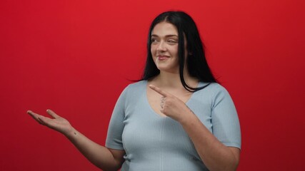 Young woman with long hair and a gray top gestures towards her palm against a vibrant red background, emphasizing a concept with a confident smile.