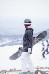 A girl with a snowboard in her hands walks along the top of a mountain, wearing a helmet and yellow glasses