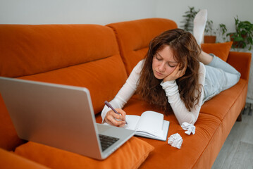 Young woman lies on an orange sofa, her head resting on her hand, as she tries to write in a notebook with crumpled paper beside her, suggesting a creative block, while a laptop sits nearby