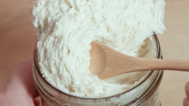 Stretchy wheat sourdough levito madre being scooped with a wooden spoon from a glass jar, showing airy texture and gluten development in a close-up view