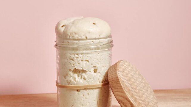 Wheat sourdough levito madre rising in time-lapse with a domed top forming above the rim of the glass jar as fermentation pushes the dough upward, wooden lid placed aside