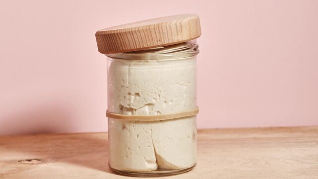 Wheat sourdough levito madre gradually rising during time-lapse inside a glass jar with loose wooden lid, early fermentation stage with soft texture and small bubbles forming