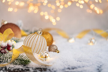 White and gold ornaments with glitter ribbon and lights rest on snow, creating a festive and...