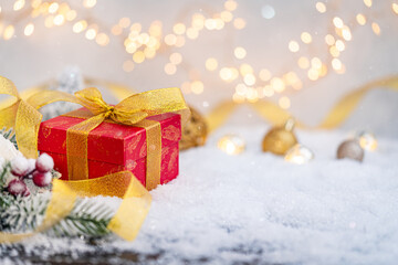 Festive red gift box wrapped in gold ribbon rests on snow with frosted pine, berries, and glowing lights in the background, creating a cozy holiday atmosphere. Ideal for holiday greeting cards,