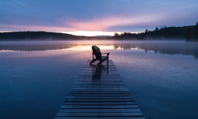 Tranquil morning on a lake. A wooden Adirondack chair sits on a pier extending into a calm lake at sunrise.  Misty morning light reflects on the water