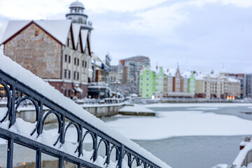 A winter scene along a snowy riverfront, featuring a decorative iron railing dusted with snow and a row of colorful buildings of Rybnaya Derevnya (Targ Rybny), in the defocused background.