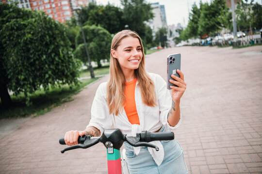 Young woman on an electric scooter smiling while taking a selfie in a sunny city park outdoor urban scene