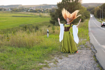Young red-haired woman in a vintage green classic dress standing in a field near the road