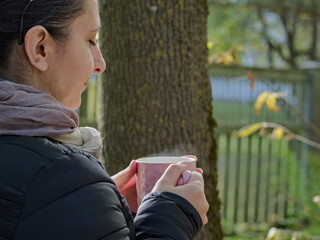 Fototapeta premium A woman in an autumn park with a drink - Woman with a Mug Leaning Against a Tree in an Autumn Park - Person Enjoying a Quiet and Peaceful Moment with a Warm Drink