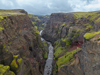 Aerial view of Icelandic canyon with river, mossy cliffs, volcanic rock, and moody sky.

