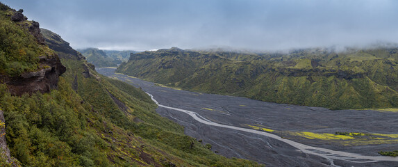 Braided river valley with volcanic sand, lush green slopes, and misty Icelandic mountains.