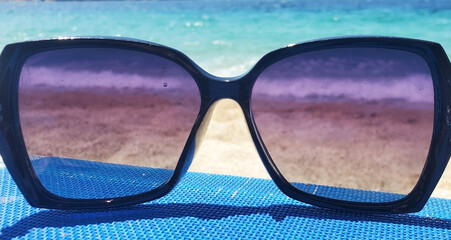 A pair of dark-framed sunglasses rests on a blue beach chair by the sea. A sandy beach and turquoise water with gentle waves are visible in the background. A sunny day, relaxation, and carefree mood.