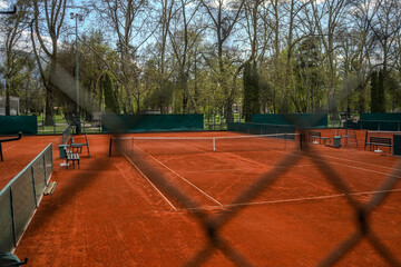 Naklejka premium Fenced tennis court in a wooded setting. The image is taken from behind a chain link fence with black noise on the court.The tennis court has a natural, clay-red colored surface with a white line.