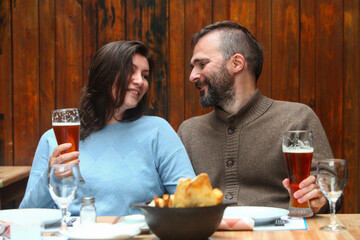 Cheerful middle-aged couple in a rustic restaurant enjoying drinks together, smiling and talking over tall beer glasses.