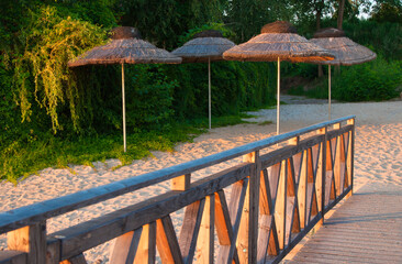 Słoneczna plaża z drewnianą balustradą, piaszczystym brzegiem i rustykalnymi słomianymi parasolami na tle bujnej zieleni. Idealne do zdjęć podróży, wakacji i relaksu na świeżym powietrzu.