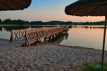 Spokojny widok plaży o zachodzie słońca, z długim drewnianym molo nad spokojną wodą, piaszczystym brzegiem i słomianymi parasolami, przywodzący na myśl relaks i spokojne chwile na świeżym powietrzu.