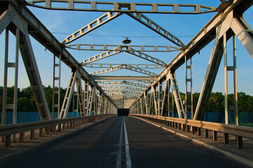 Nowa Sól - Stalowy most kratownicowy rozciąga się nad rzeką Odrą w ciągu drogi nr 315, otoczony niebieskimi barierkami i bujnymi drzewami. Ciepłe światło podkreśla detale konstrukcyjne. © Pol Badger