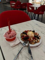 Plate with waffle, ice cream, strawberries, and cookies next to a cup of strawberry ice cream on a marble table with red chairs.