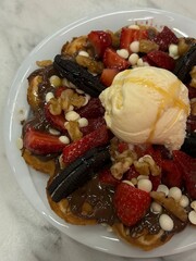 Close-up of a waffle topped with vanilla ice cream, strawberries, chocolate, cookies, nuts, and white chocolate pieces on a white plate.
