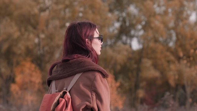 Teen girl standing with her back to the camera, turning around with a gentle smile, wearing sunglasses and a warm brown coat in an autumn landscape.