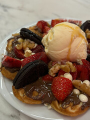 Close-up of a waffle topped with vanilla ice cream, strawberries, chocolate, cookies, nuts, and white chocolate on a white plate.