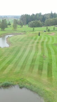 Aerial footage of a golf course with visible fairways, sand bunkers, ponds, and golfers playing under cloudy skies in a scenic countryside landscape.
