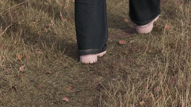 Close-up of legs in dark jeans and white boots, walking away and then running towards the camera on a grassy path with autumn leaves.