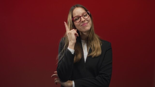 Woman pointing index finger to camera in red studio with glasses and blazer; confidence assertiveness charm.