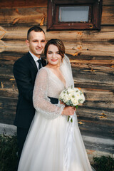 Bride in an elegant white gown and groom in a classic suit pose by a rustic wooden wall. She holds a bouquet of white flowers. A warm atmosphere of love, style, and intimate romance.