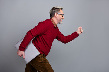 Confident middle-aged businessman in casual attire walking with a laptop against a gray background