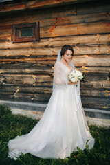 Bride in white dress holding bouquet near rustic wooden wall. Her veil flows in the wind, creating a soft, romantic, countryside atmosphere.