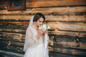 Bride in white dress holding bouquet near rustic wooden wall. Her veil flows in the wind, creating a soft, romantic, countryside atmosphere.