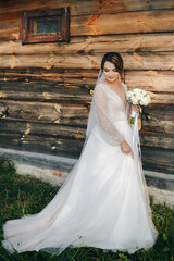 Bride in white dress holding bouquet near rustic wooden wall. Her veil flows in the wind, creating a soft, romantic, countryside atmosphere.