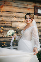 Bride in white dress holding bouquet near rustic wooden wall. Her veil flows in the wind, creating a soft, romantic, countryside atmosphere.