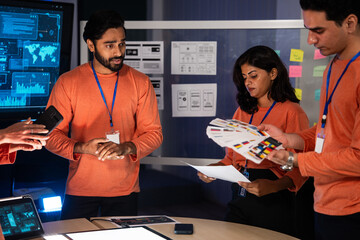 A diverse creative team is collaborating on a critical UI, UX design problem late at night in a tech office. Focused on color palettes and wireframes for a new mobile application. Project meeting