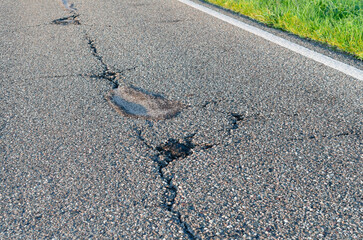 German Road Cracks in Late Sunlight; Pothole with roadside greenery and road marking in a diagonally road view