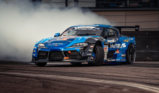 Latvia, Riga 03-05.10.2025. FIA Drifting cup,  a blue Toyota Supra drifting on a track with smoke billowing out of it, surrounded by a fence and a wall in the background